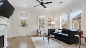 A spacious living room with a fireplace, a ceiling fan, a black leather sofa, a coffee table, and a large window. - Luxury Cottage in Historic Beauregard Town  (Baton Rouge)