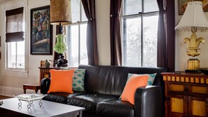 Una habitación con un sofá de cuero negro, dos cojines naranja, un jarrón verde y una lámpara blanca sobre una mesita de madera.