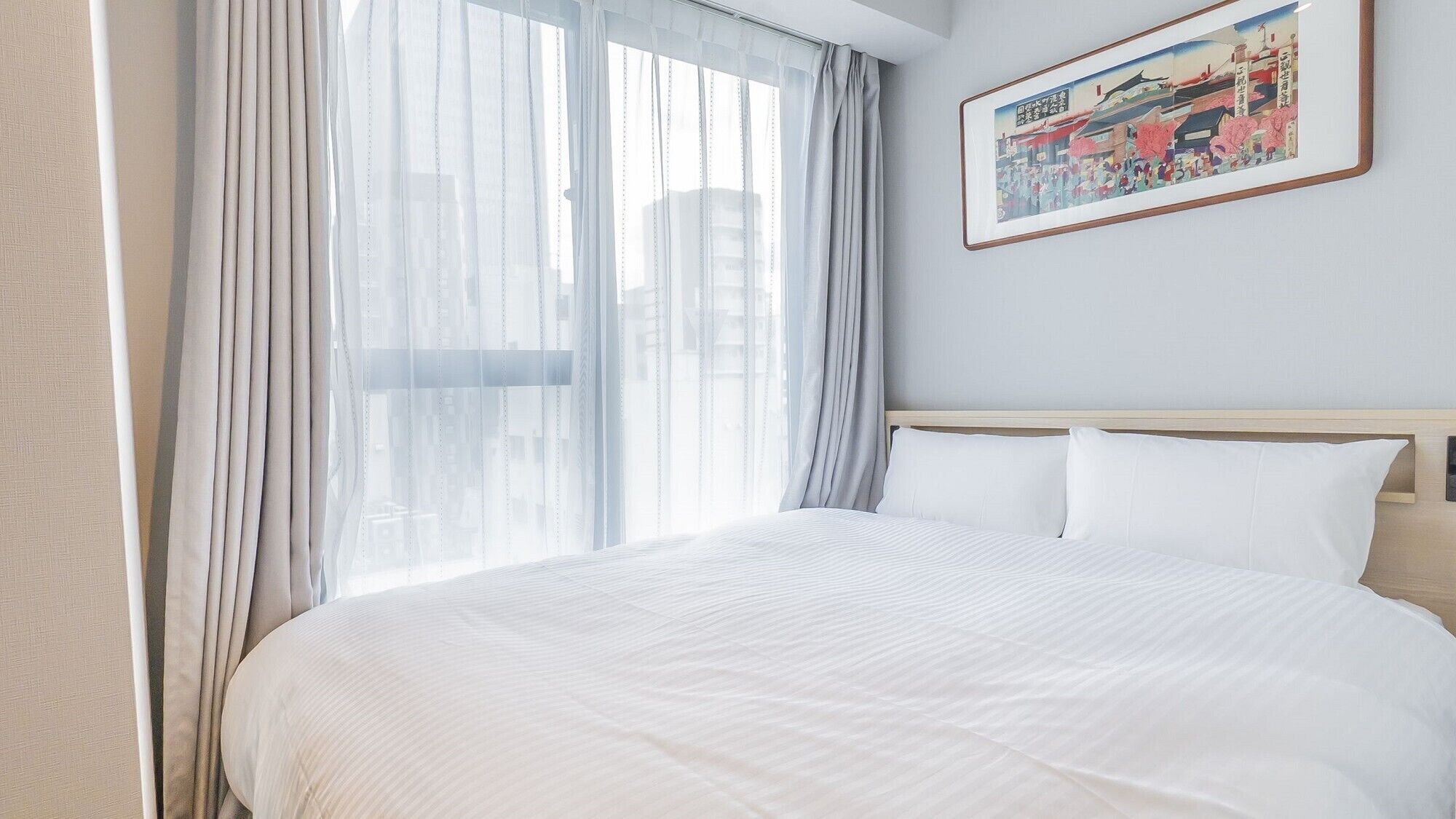 A hotel room with a large bed, sheer curtains, a framed picture on the wall, and a view of buildings outside.