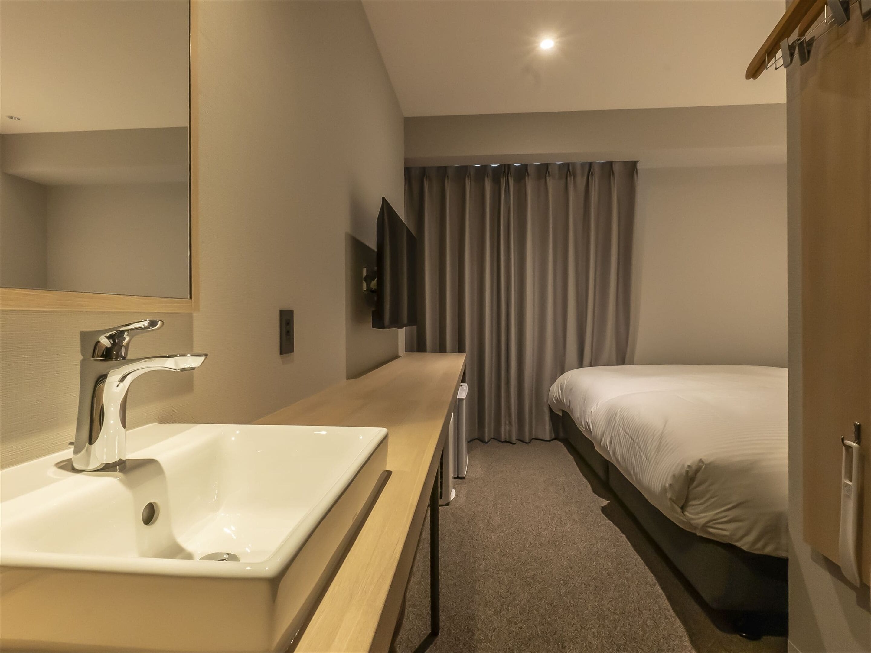 A modern hotel room with a large mirror, a white sink, a flat-screen TV mounted on the wall, a bed with white bedding, and a window with grey curtains.