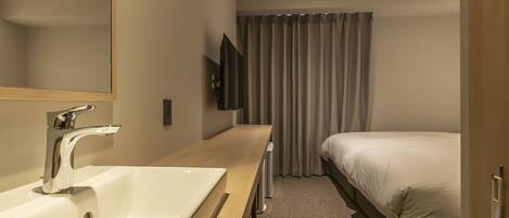 A modern hotel room with a large mirror, a white sink, a flat-screen TV mounted on the wall, a bed with white bedding, and a window with grey curtains.