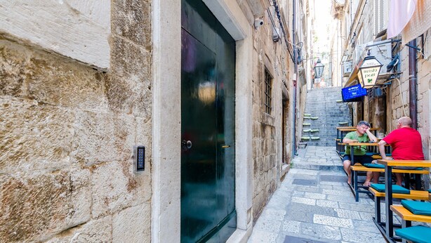 A narrow alley with stone walls, a dark door, and outdoor seating with tables and chairs.