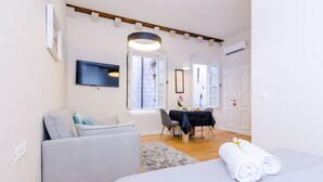 A modern hotel room with a bed, a sofa, a dining table with chairs, and a TV mounted on the wall.