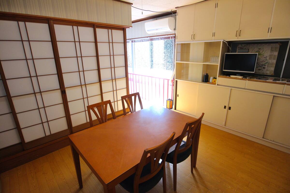 A traditional Japanese-style room with tatami flooring, a wooden dining table with chairs, a sliding shoji screen, and a TV on a cabinet.