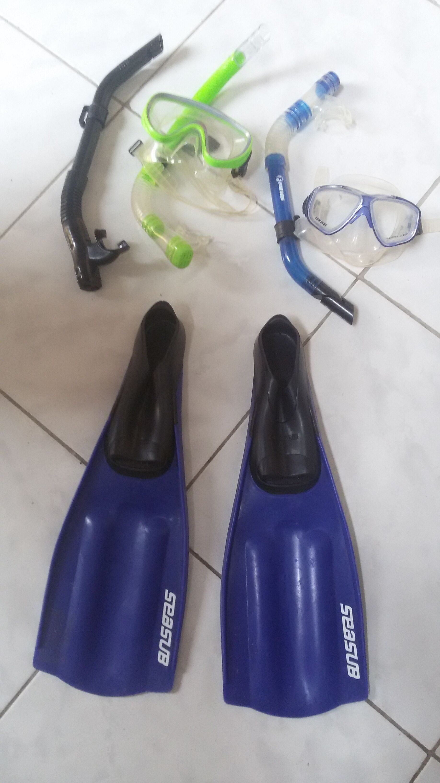 Un par de aletas de snorkel azules, un snorkel verde, una botella de plástico transparente, una máscara de snorkel azul y un tubo de snorkel negro sobre un piso de baldosas.