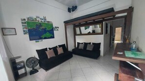 Una habitación con un sofá negro, un ventilador de pie, un mapa en la pared y un espejo sobre una estructura de madera.