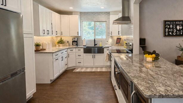 A modern kitchen with white cabinetry, a stainless steel refrigerator, a granite countertop, and a window with a view of trees.