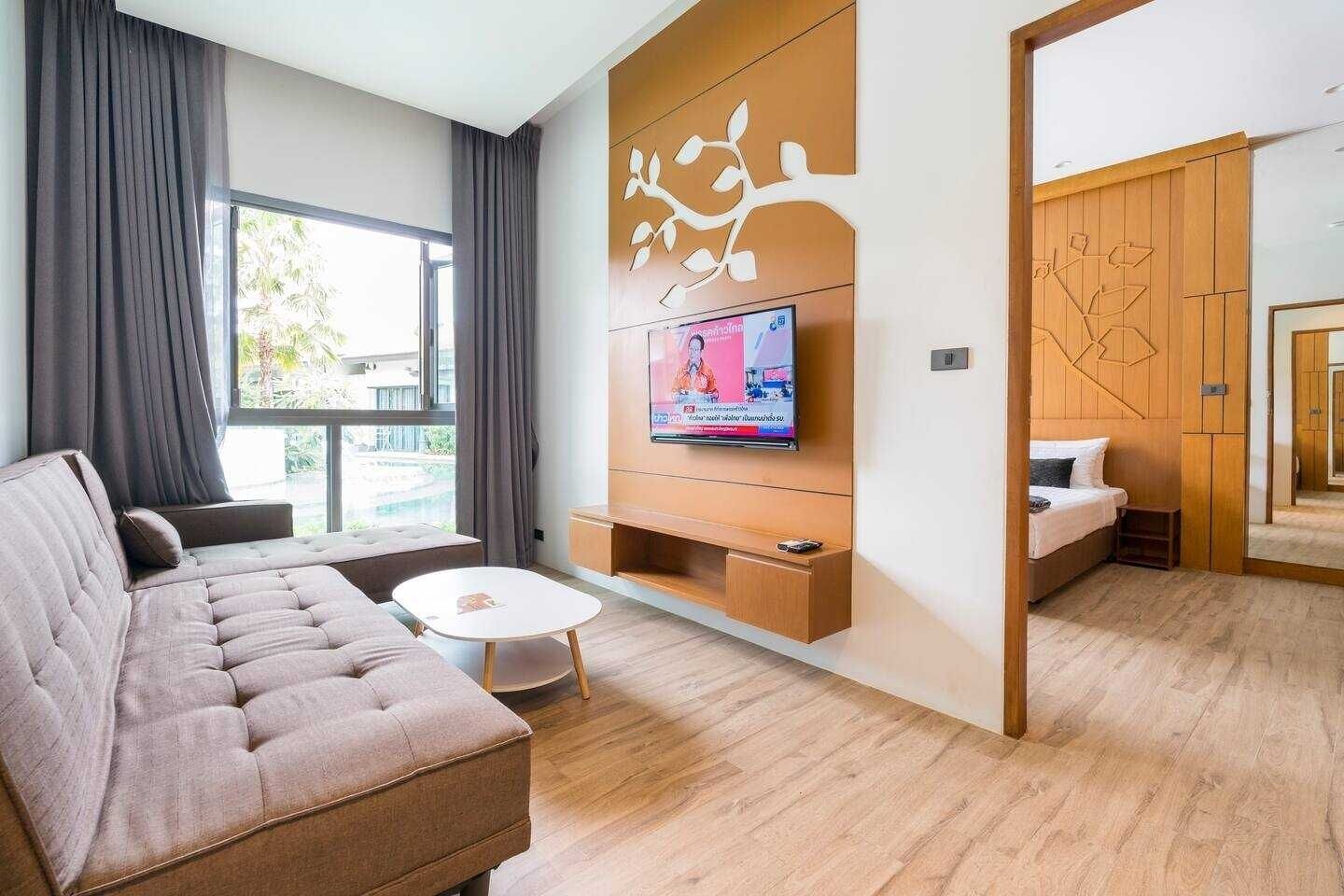 A modern living room with a sofa, a coffee table, a TV mounted on a wooden panel, and a bedroom visible through an open door.
