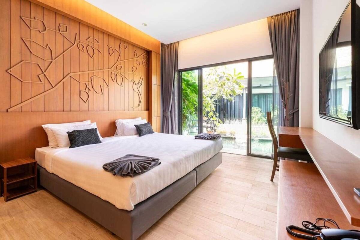 A modern hotel room with a large bed, a wooden headboard with a unique design, a desk, and a large window offering a view of greenery.