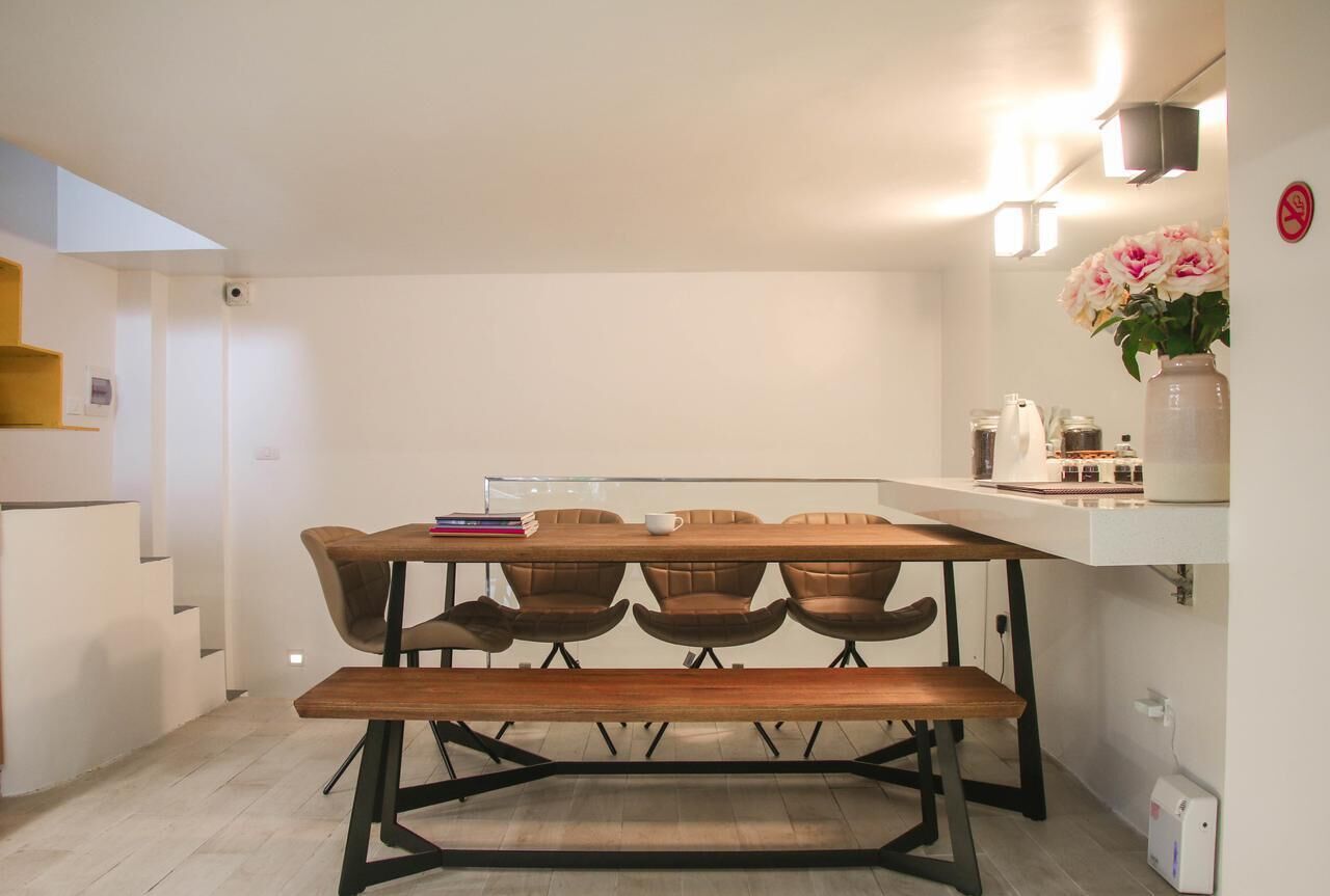 A modern kitchen with a wooden table, four chairs, and a bench. There is a white countertop with a vase of flowers and various kitchen items.