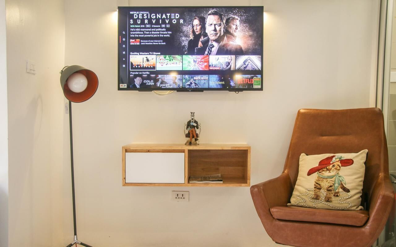 A living room with a mounted TV displaying a show, a wooden shelf with a figurine, a brown leather armchair with a pillow, and a floor lamp.