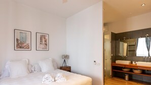 Une chambre d’hôtel comprenant un lit, deux tableaux encadrés, une table de chevet avec une lampe, un plancher en bois et une salle de bain avec un grand miroir, un lavabo et un porte-serviettes.