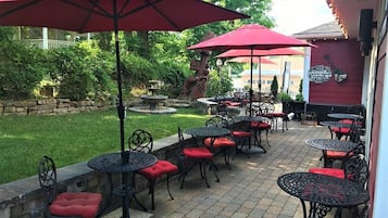 An outdoor patio area with multiple tables and chairs, red umbrellas, and a stone wall.
