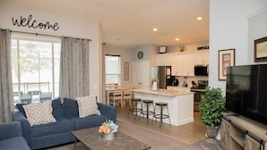 A modern living room with a blue sofa, a coffee table with flowers, and a kitchen area with white cabinets and a large TV.