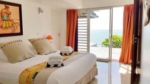 Una camera d'hotel con un letto, cuscini, una lampada, un quadro e vista sul mare tramite una porta scorrevole.