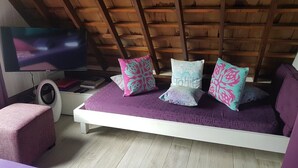 Una camera con soffitto spiovente, un letto con materasso viola, due cuscini, una televisione e un piccolo tavolo bianco con libri e un altoparlante.