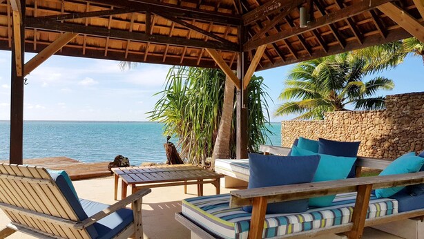 Zona salotto coperta all'aperto con mobili e cuscini in legno, affacciata su una spiaggia con palme e l'oceano.