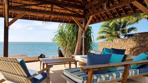 A covered outdoor seating area with wooden furniture and cushions, overlooking a beach with palm trees and the ocean. - Villa Vaihau - Vacation rental by the sea (Papeotai)