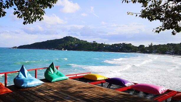 A wooden deck with colorful cushions overlooking a beach with waves and a hill in the distance. - So Good Haad Rin Hostel (Ko Pha-ngan)
