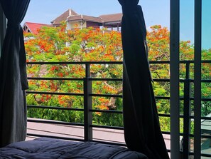 A room with a bed, a window with curtains, and a view of a building with red-roofed houses and a tree with red flowers.