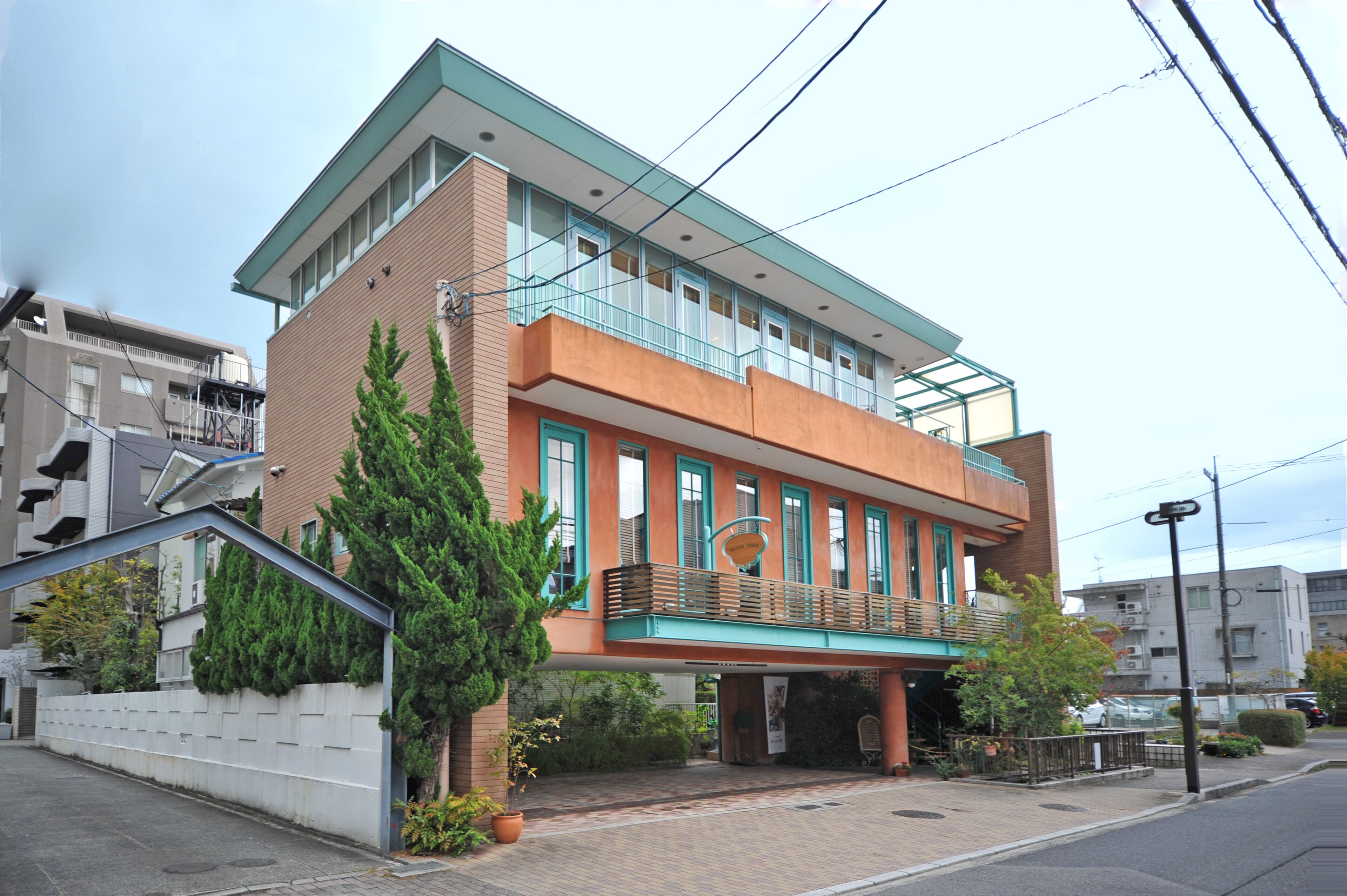 A two-story building with a flat roof, a balcony on the second floor, and a small entrance area with a potted plant.