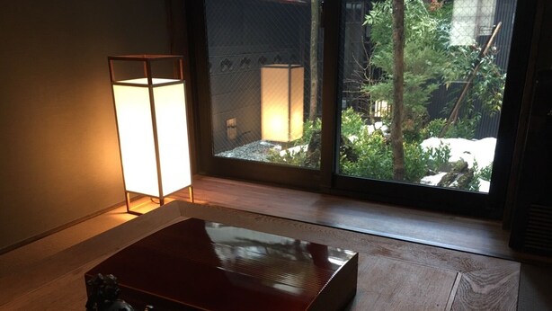 A room with a wooden floor, a dark red table, a small statue, and a large window with a view of greenery and a building. - Ichii no Niwa (Takayama)