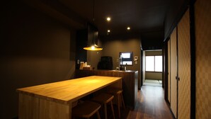 A modern kitchen with a wooden island and bar stools, a hanging pendant light, and a view into the dining area. - Ichii no Niwa (Takayama)