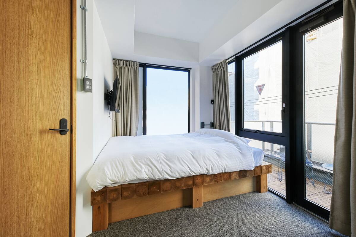 Une chambre avec un lit en bois, une grande fenêtre et une porte coulissante donnant sur un balcon extérieur.