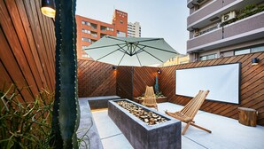 Un espace extérieur aménagé avec un foyer, des fauteuils de jardin en bois et un grand parasol.