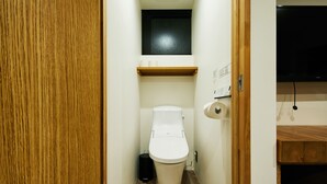Ein kleines, schmales Badezimmer mit Toilette, einem Holzregal darüber und einer schwarzen Mülltonne neben der Toilette.