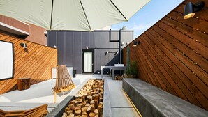 Ein moderner Außenbereich mit Holzbank, Feuerstelle und einem großen Sonnenschirm, der Schatten spendet.
