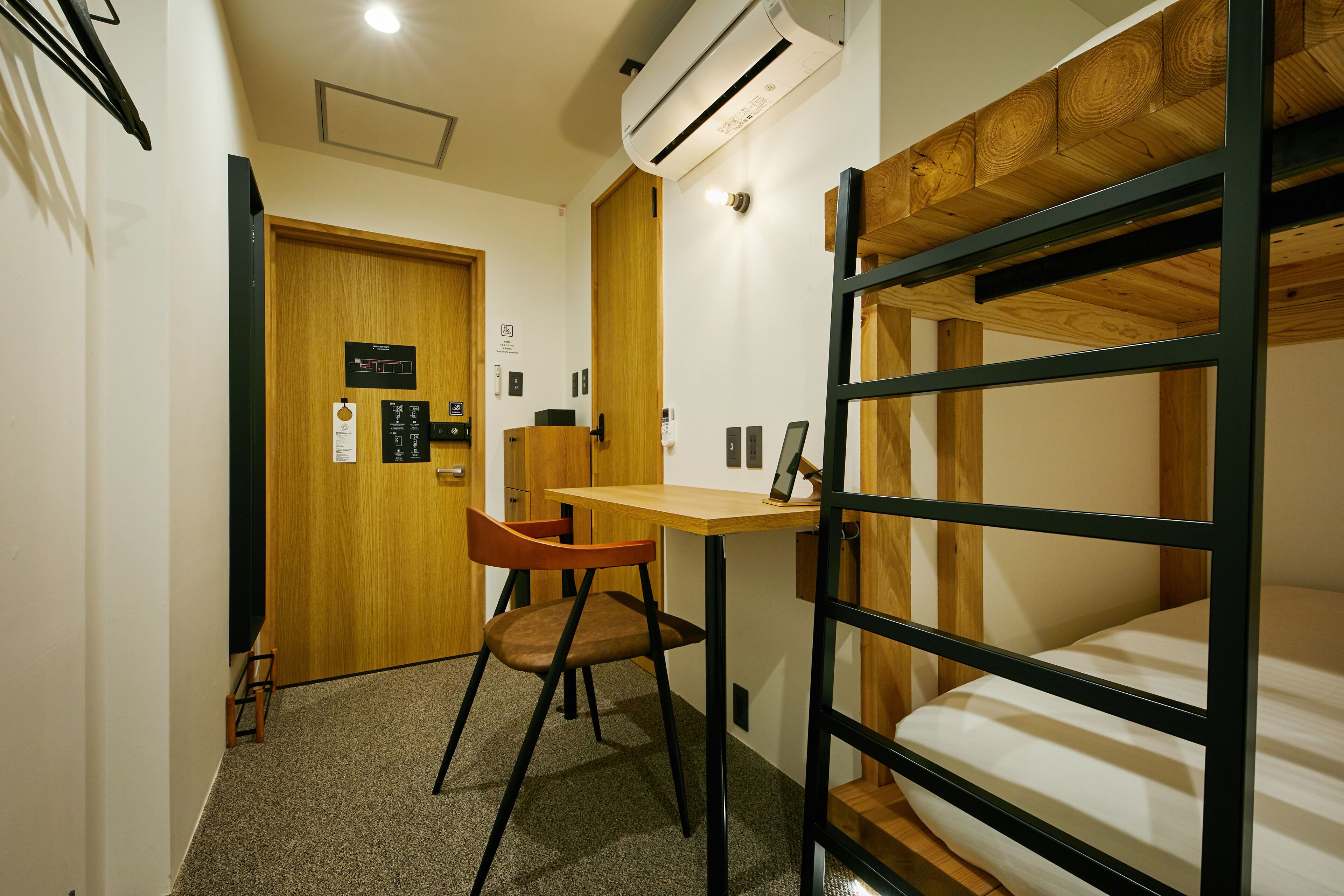 A compact room with a bunk bed, a desk with a chair, a wooden door, and an air conditioning unit.
