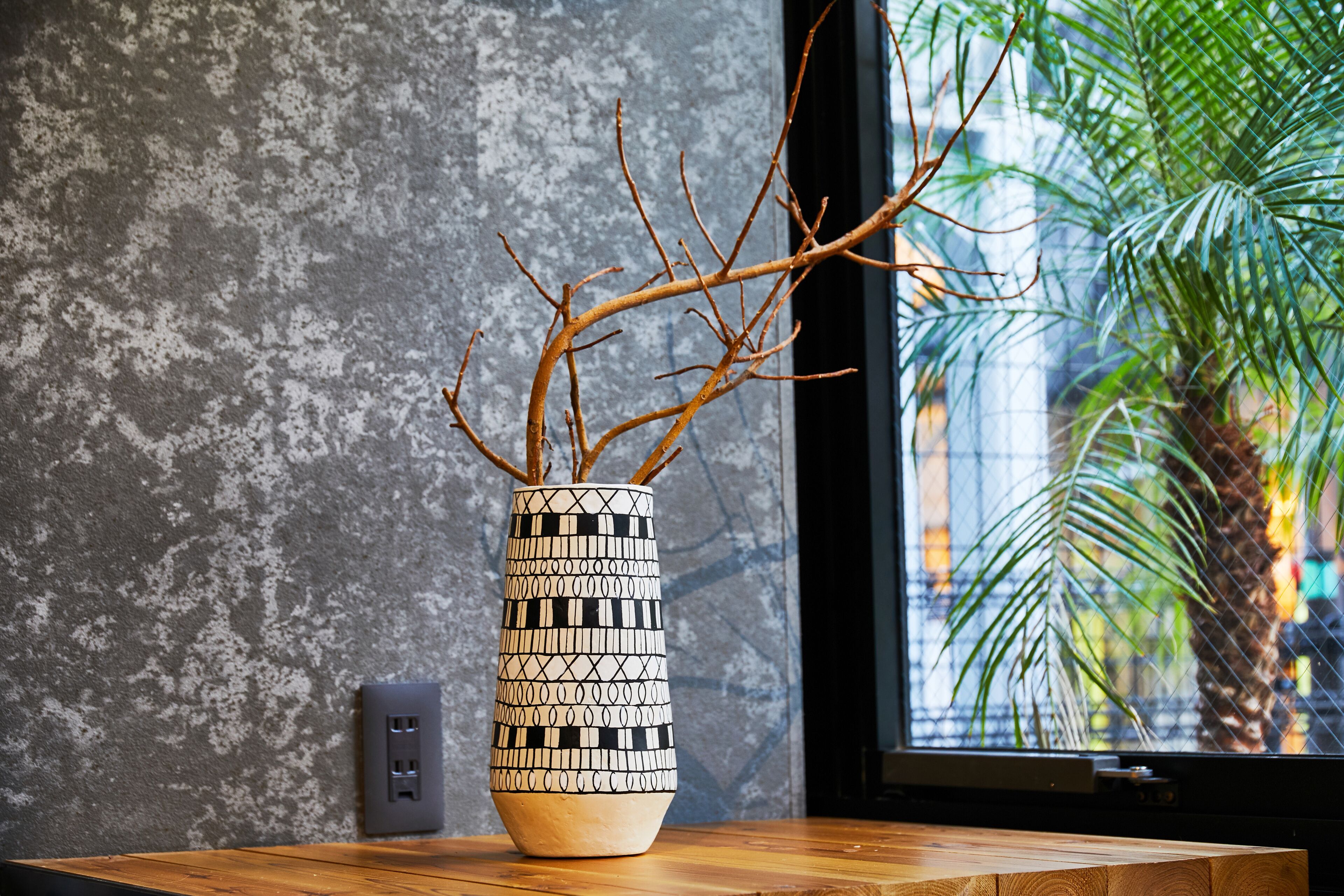 A vase with a geometric pattern on a wooden table next to a textured wall and a window with a view of a palm tree.