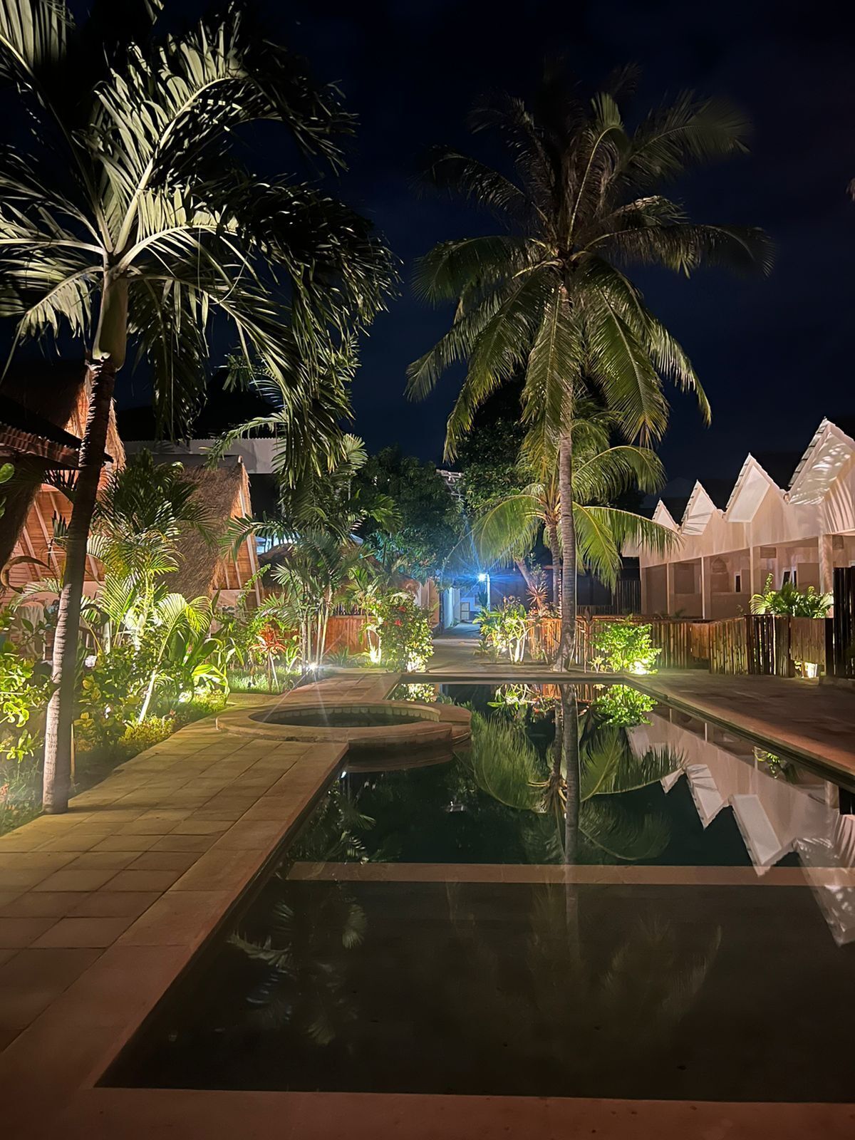 A nighttime resort pool area with palm trees, a clear pool, and overwater bungalows.