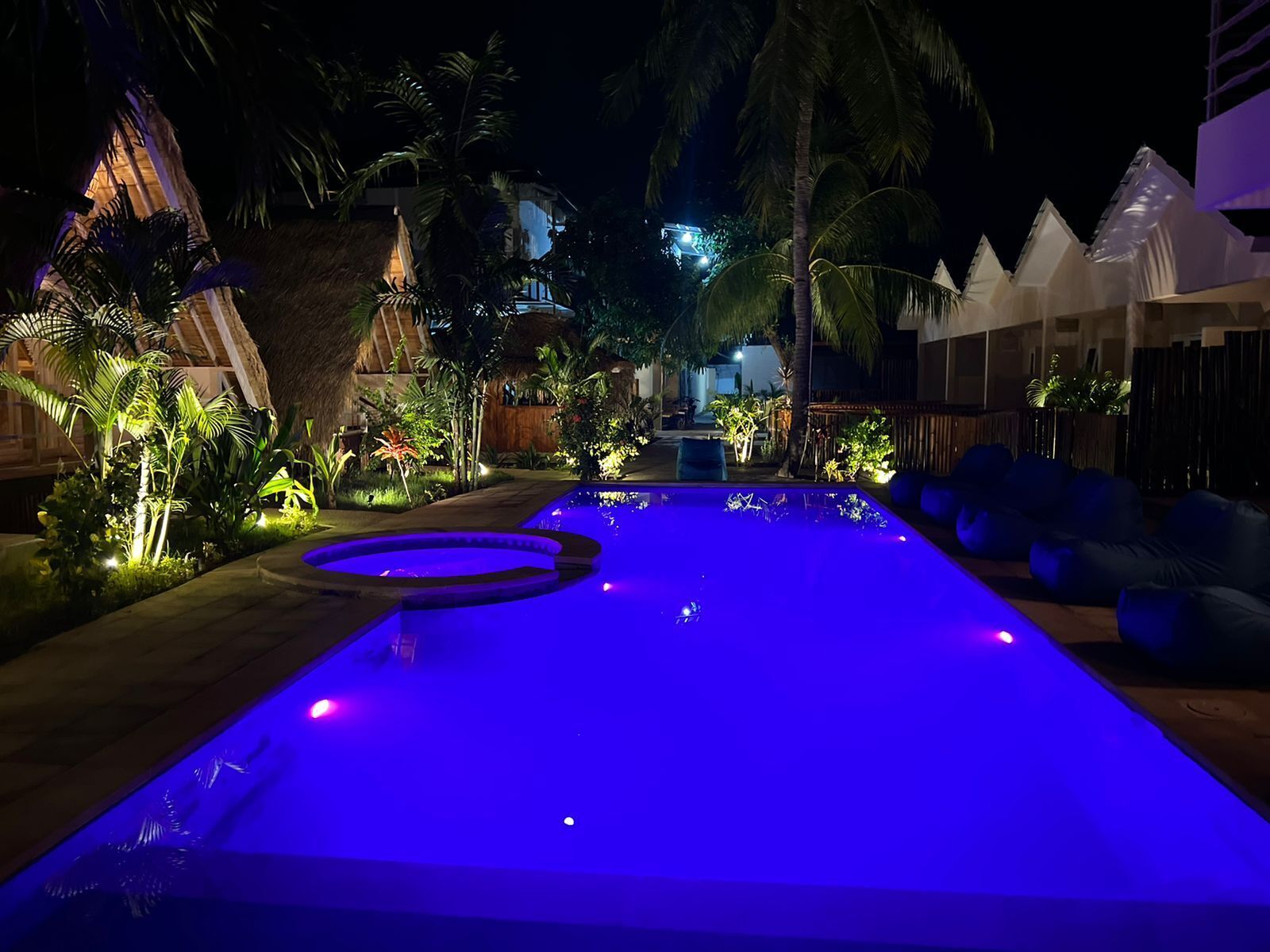 Une piscine dont l’eau est illuminée de couleurs bleues, entourée de plantes tropicales et bordée de constructions aux toits de chaume.