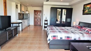 Ein Schlafzimmer mit Bett, Nachttisch und Fernseher. Es gibt eine Küchenzeile mit Schränken und einer Mikrowelle.