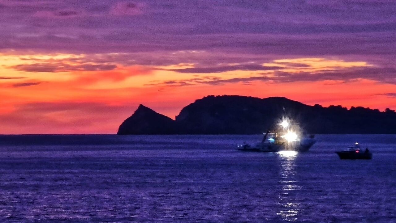 A sunset over a body of water with boats and a distant mountain.