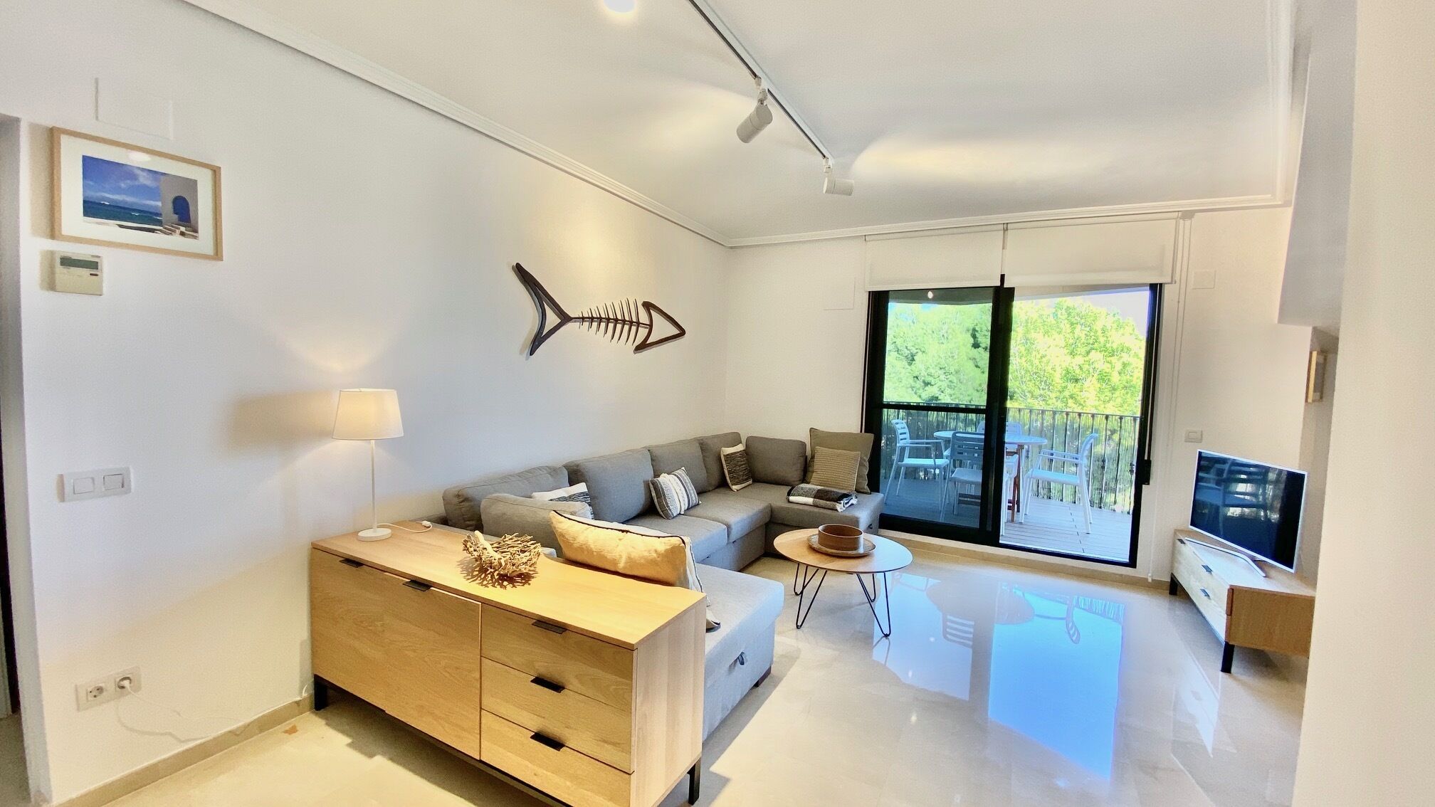 Un salon moderne avec un canapé, une table basse et un téléviseur. Une grande fenêtre offre une vue sur les arbres, et il y a un balcon.