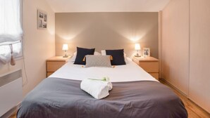 Quarto de hotel com uma cama grande, duas mesas de cabeceira com luminárias, uma janela com cortinas e um radiador.
