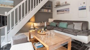Uma sala de estar moderna com um sofá cinza, uma mesa de centro de madeira com copos de vinho, uma escada com guarda-corpo branco e uma estante com livros e objetos decorativos.