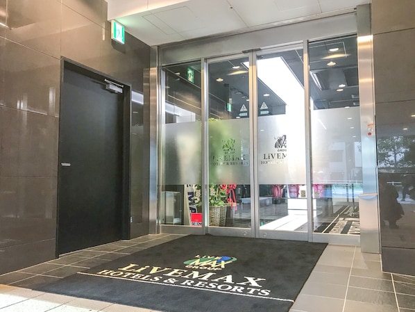 L’entrée d’un hôtel, avec une porte noire et un hall d’entrée en verre sur lequel on peut lire les inscriptions « LIVEMAX GROUP » et « HÔTELS ET RESORTS ».