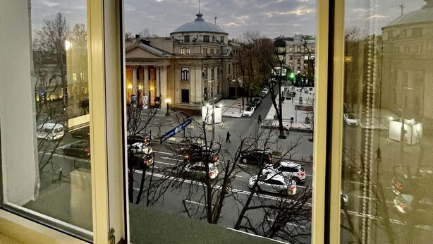 Blick durch ein Fenster auf ein Gebäude mit einer Kuppel, einen Parkplatz und eine Straße mit Autos und kahl stehenden Bäumen.