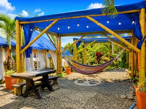 Un espacio al aire libre cubierto con una mesa de madera y bancos, rodeado de estructuras de bambú y lonas azules.