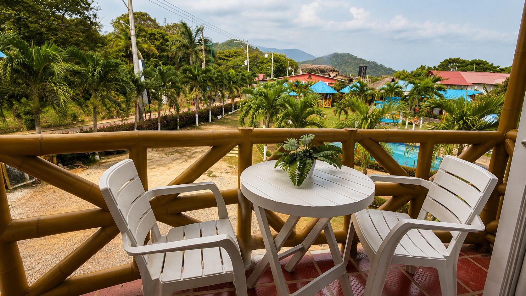 A balcony with two white plastic chairs and a small round table, overlooking a beach and a pool area.