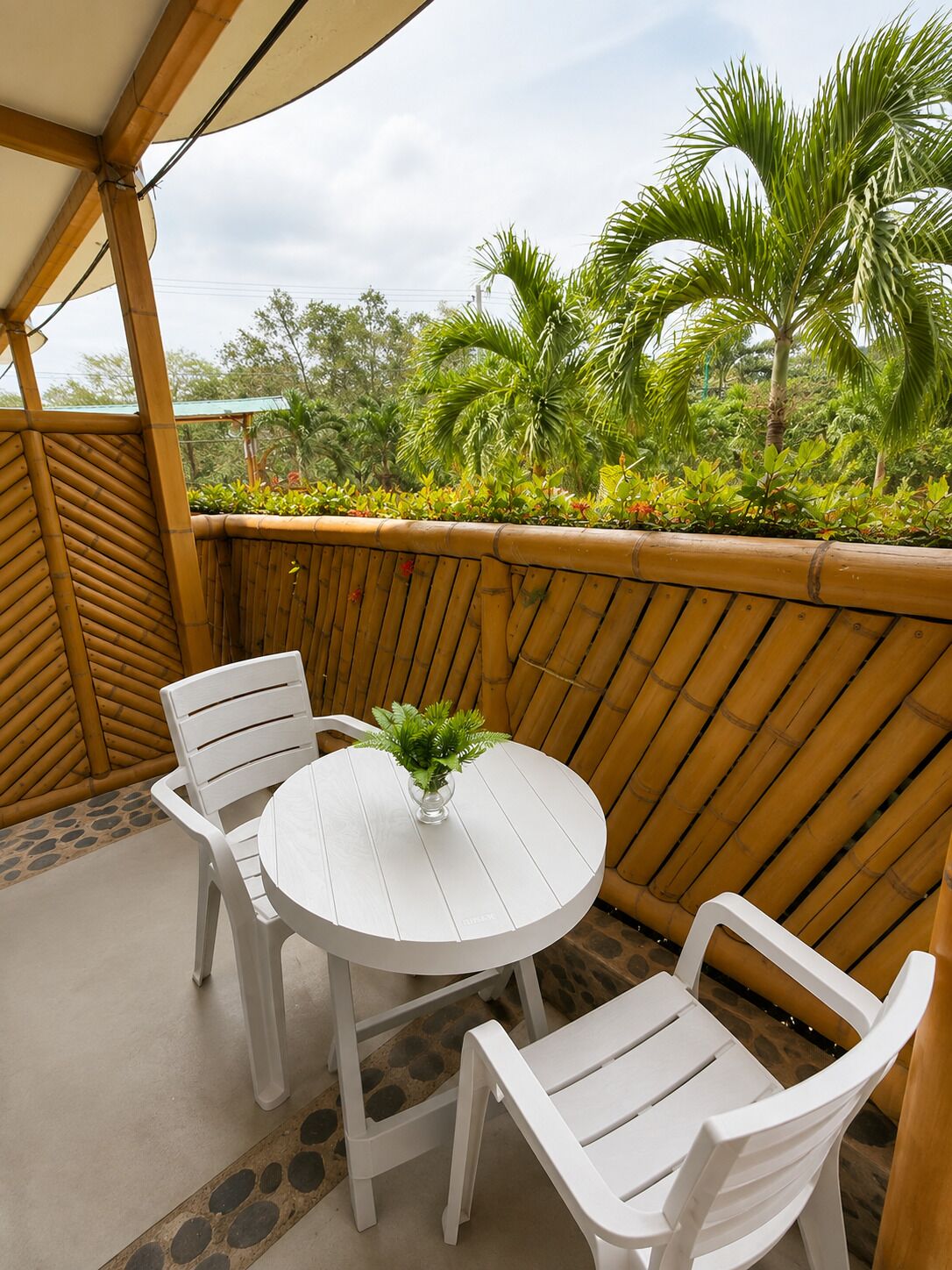 A covered outdoor seating area with a round table and two chairs, surrounded by a bamboo fence and tropical plants.