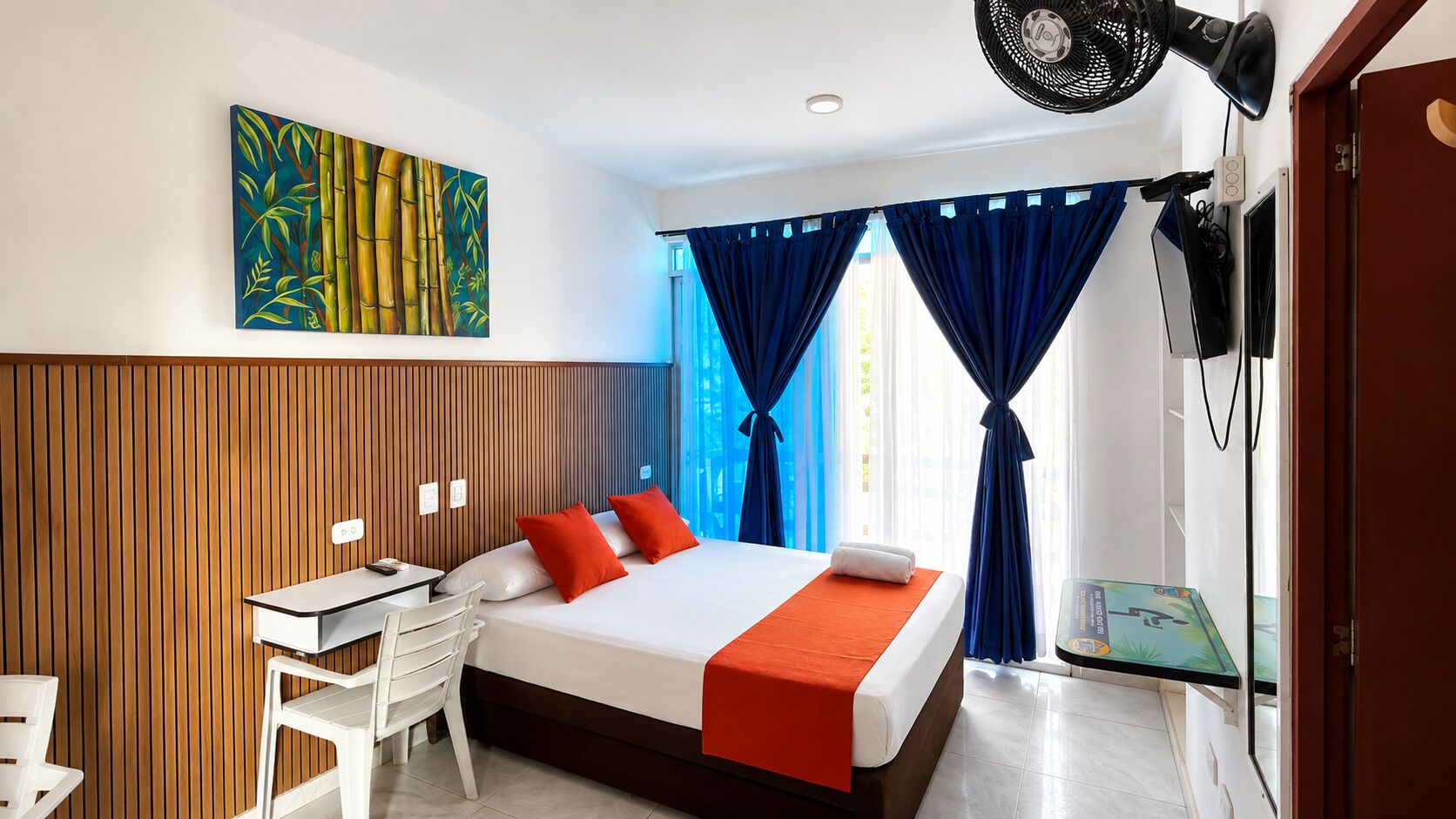 A hotel room with a bed, a desk, and a chair. There is a painting on the wall and a window with curtains.