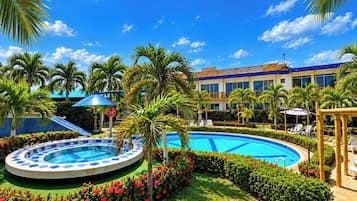 Zona de piscina de hotel con una piscina circular, palmeras y un edificio de fachada azul y blanca.