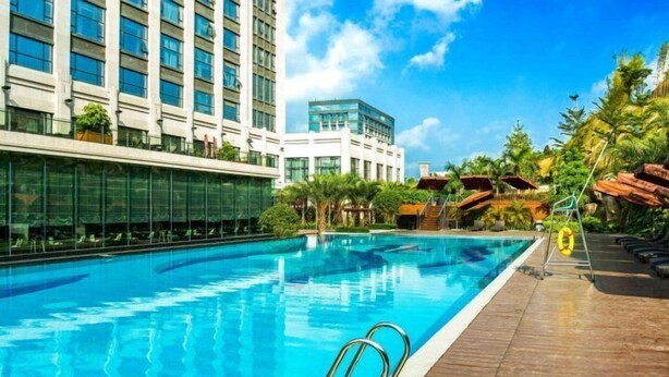 A swimming pool with a wooden deck, surrounded by palm trees and modern buildings. - Winlong International Hotel (Quanzhou)