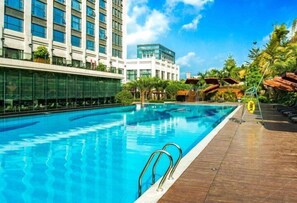 Piscina com deck de madeira, rodeada por palmeiras e edifícios modernos.