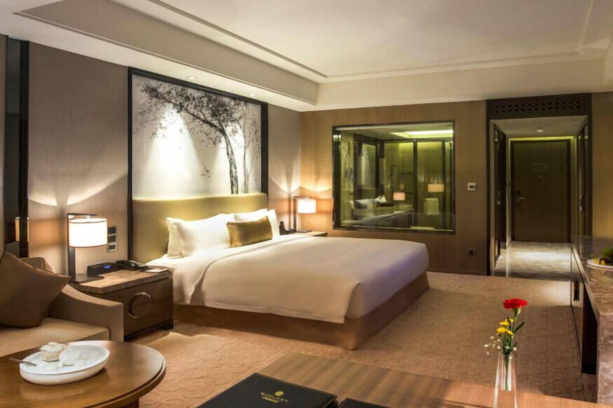 Habitación de hotel con una cama grande, mesitas de noche, un escritorio, una silla, una mesa de comedor con un jarrón de flores, un ventanal con vistas y un espejo.